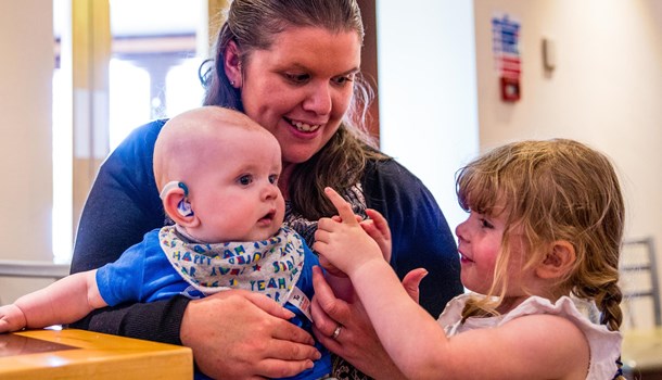 A mum smiles at her young daughter while holding a baby wearing hearing aids