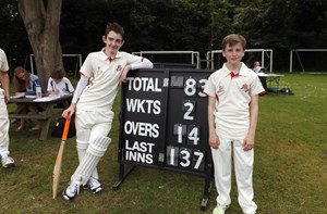 Two boys wearing cricket kit while standing next to a score board