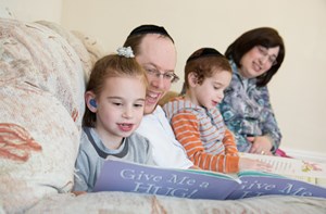 A family of four sit on a sofa watching their daughter read a book.