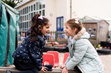 Two girls sit on a bench in a playground, smiling at each other. 