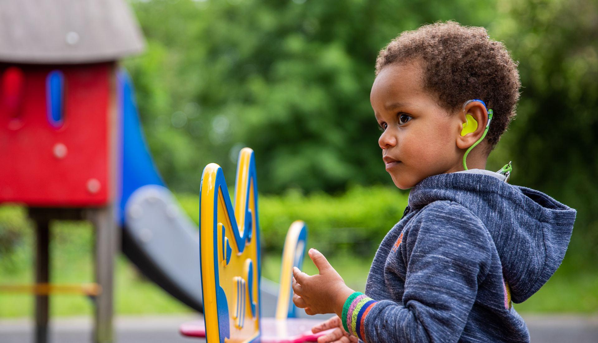 A young child with hearing aids, wearing a blue hoodie, stands in a playground near colourful play equipment.