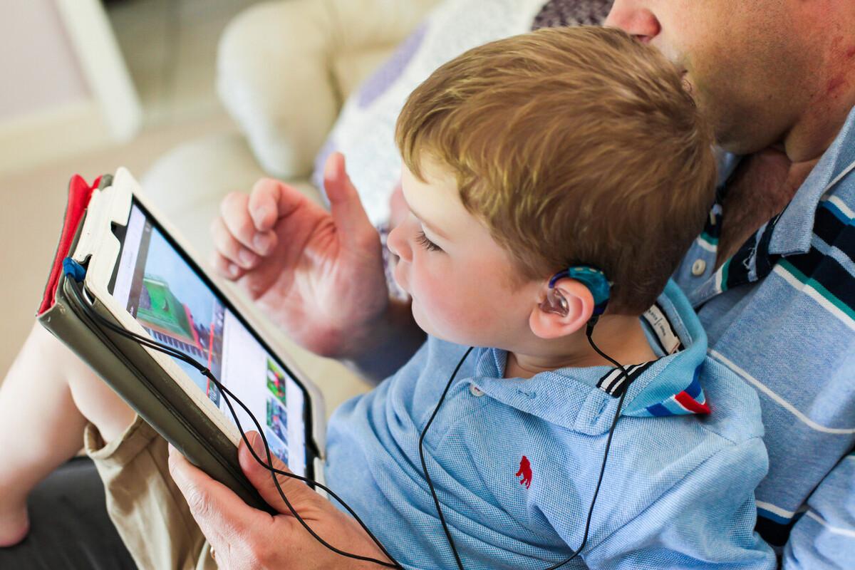 Young boy with hearing aids sits in his dad's lap watching a show on a tablet device