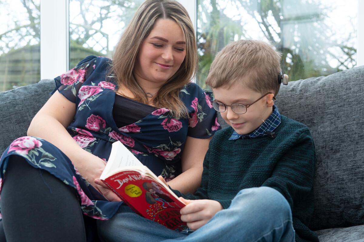A boy of about eight or nine is sat on a grey sofa, reading a book with his mum. He has blond hair and glasses. He is wearing a bone anchored hearing aid and has microtia. He is wearing a grey jumper and jeans and his mum is wearing a black and pink floral dress.