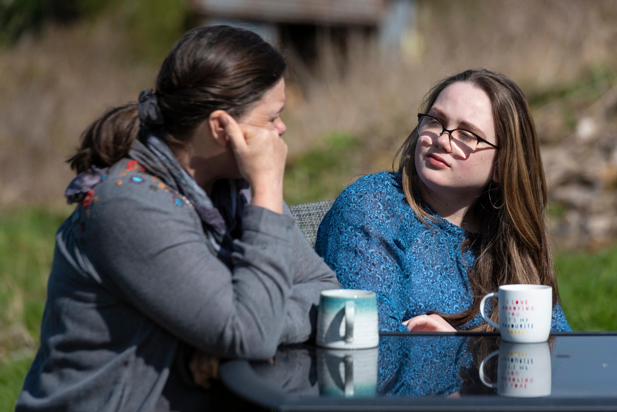 Two young women talk at a table with mugs on it