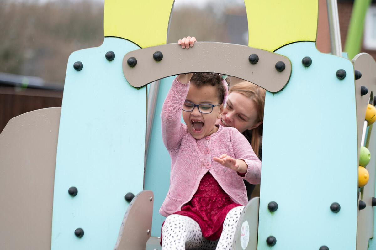 Genevieve smiling at the top of a slide in a playground with her mum behind her.