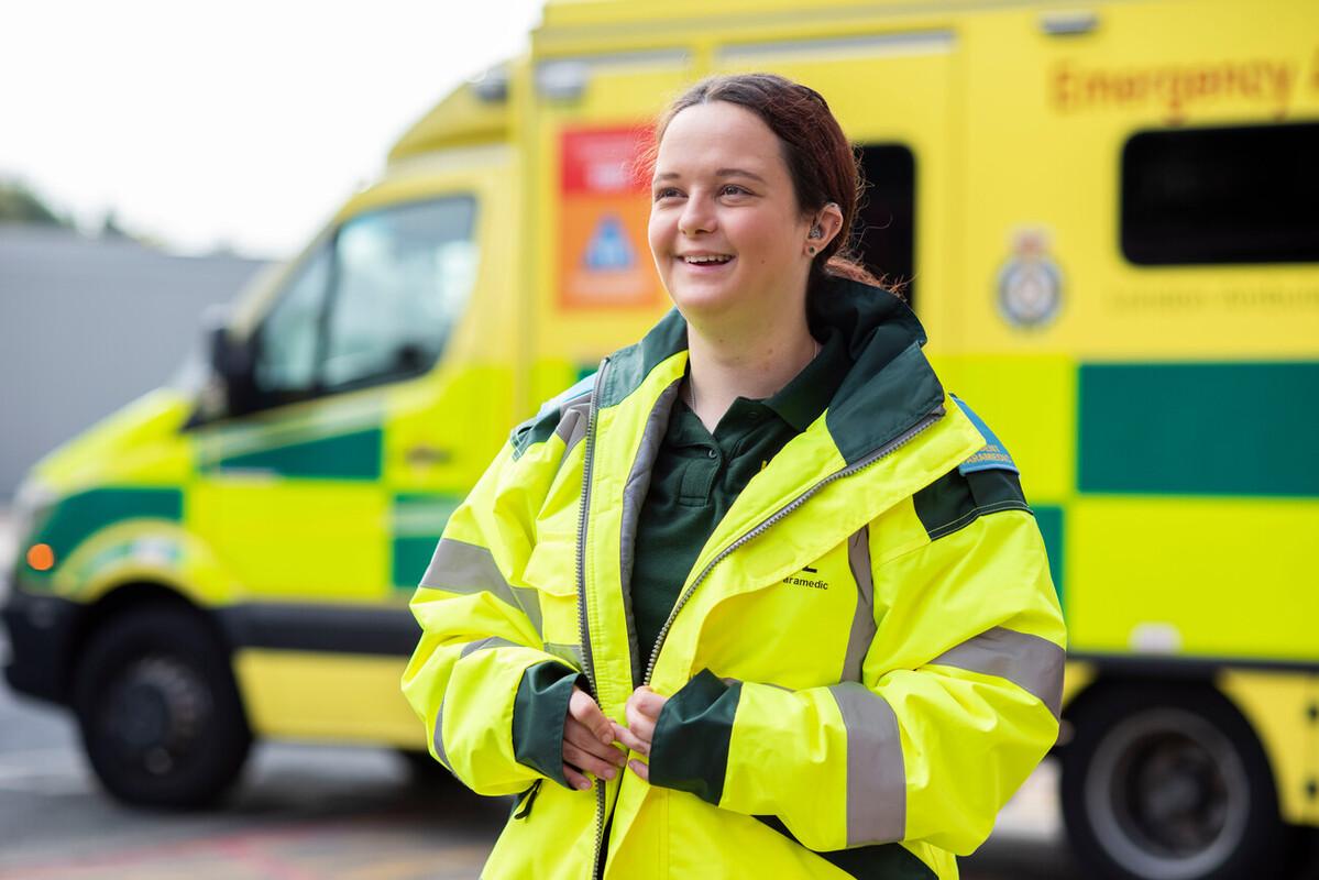 Holly smiling wearing a high-visibility paramedic coat stood in front of an ambulance.