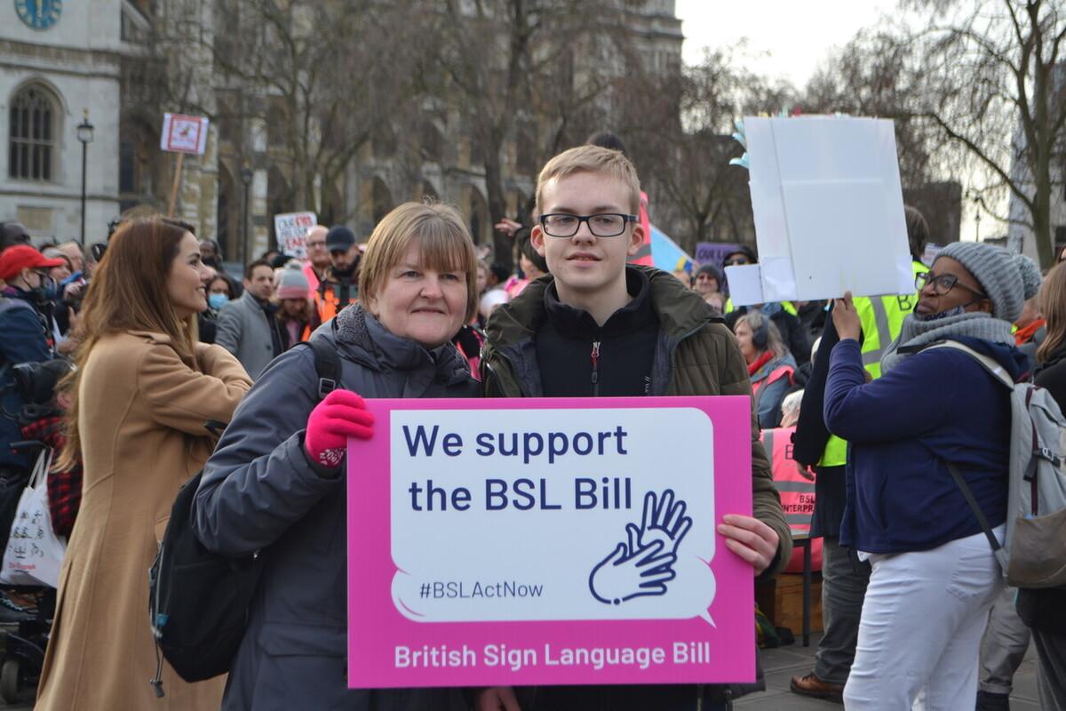 Dan and Ann standing in Parliament Square holding s white and pink sign which says "We support the BSL Bill".