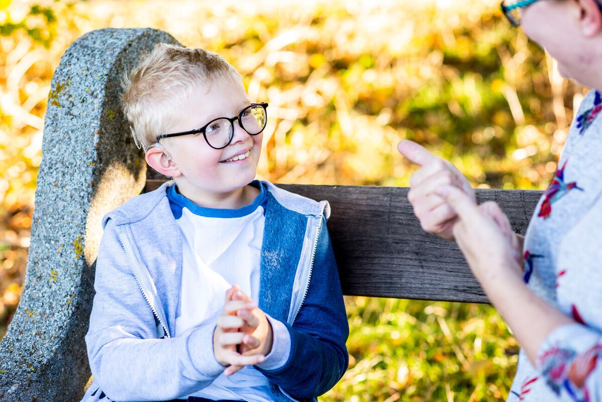 Xander sat on a park bench smiling at his mum signing.