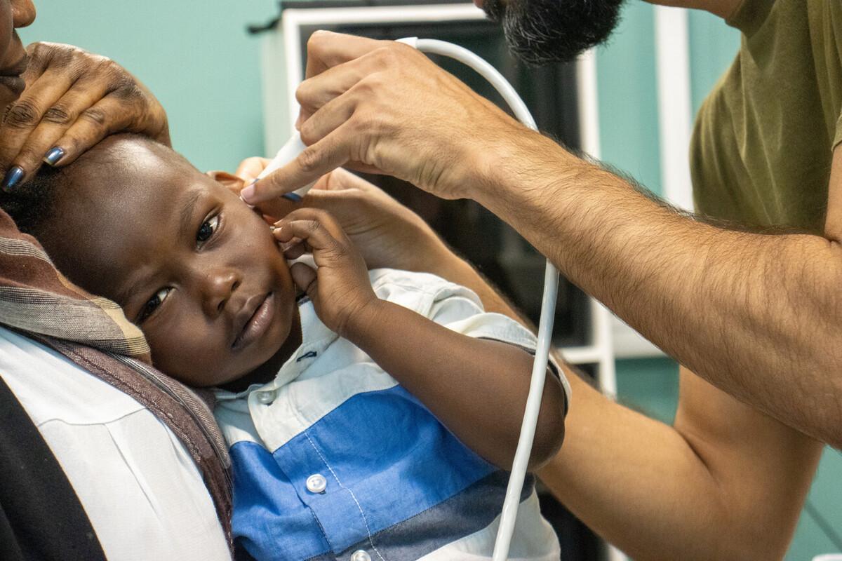 A young child is at the audiologist. He is black and is wearing a striped blue and white shirt. A caregiver is cuddling him to her chest. The doctor is examining his ear with an instrument. 