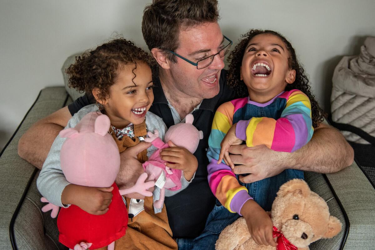 Dad Matt sitting on an armchair holding a laughing with his two daughters, Eleanor and Millie. Eleanor is holding two Peppa Pig toys and Millie is holding a teddy bear.