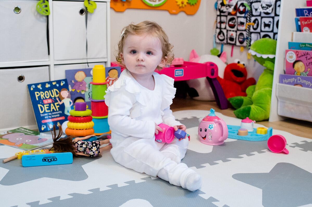 Olivia is looking at the camera and wearing white while sitting on the floor surrounded by colourful toys and a book titled 'Proud to be Deaf'.