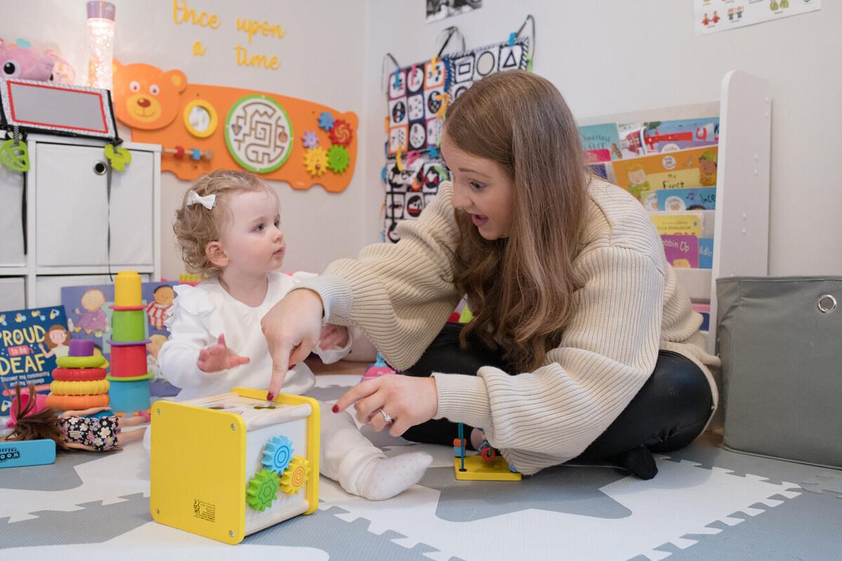 Olivia sitting on the floor looking at mum, Yasmin, who is pointing at a yellow box.