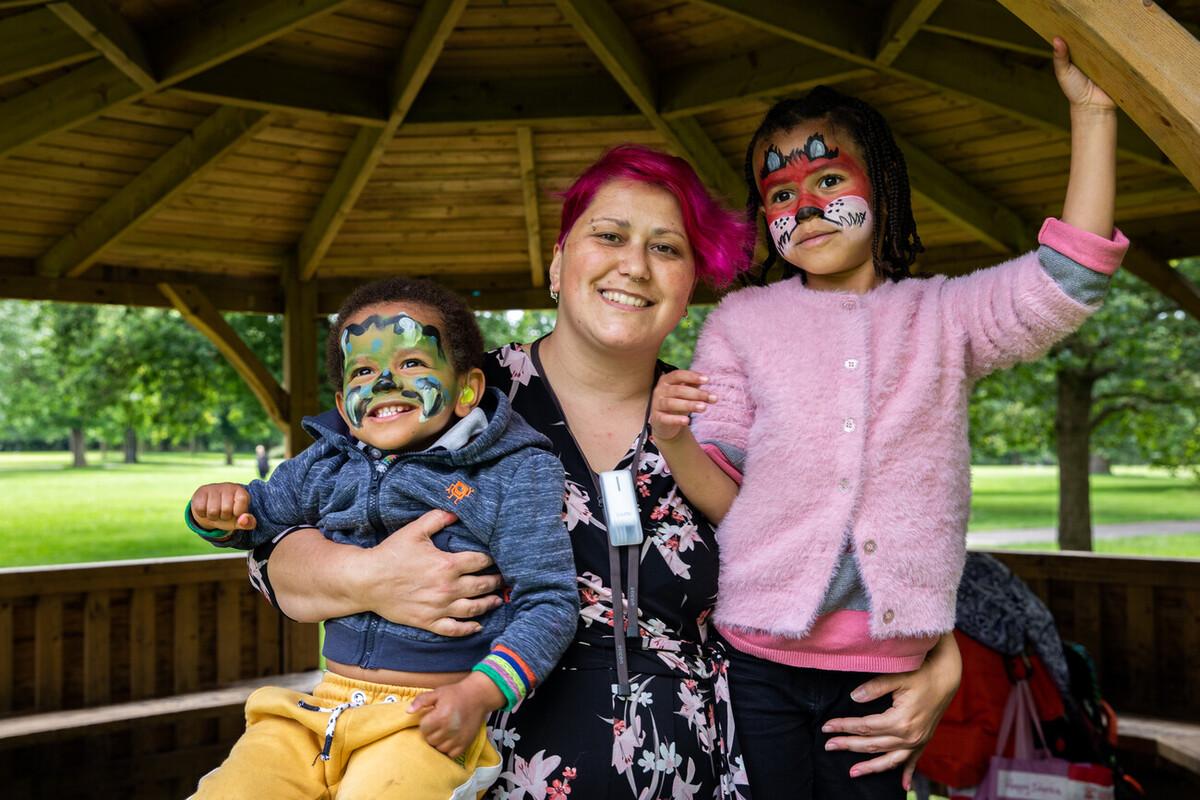 Mum Chrysanthi is stood in a bandstand in a park holding son Lamin-Phoenix and daughter Mariam-Iris, who both have their faces painted as animals.