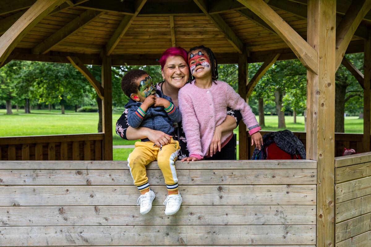 A family with a mum and two children smiling together. They are out in a park in a wooden gazebo. The children have their faces painted.