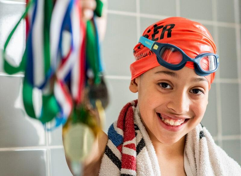 Santiago wearing a towel around his shoulders and swimming cap and goggles, holding up his swimming medals.