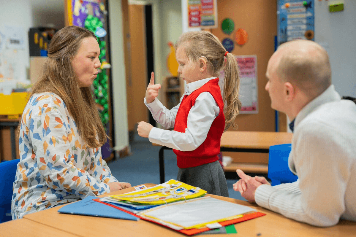 A young girl standing in a classroom signing to a woman sat on her left. A man sat on her right is watching on.
