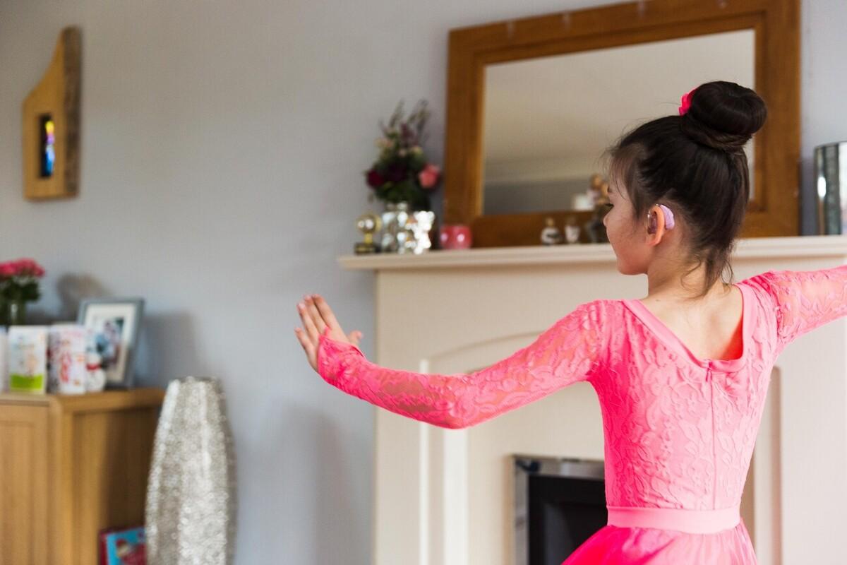 A young girl is dancing in her living room. She is wearing a hearing aid. Her hair is in a bun and she is wearing a pink dance outfit with a long skirt and lace sleeves.