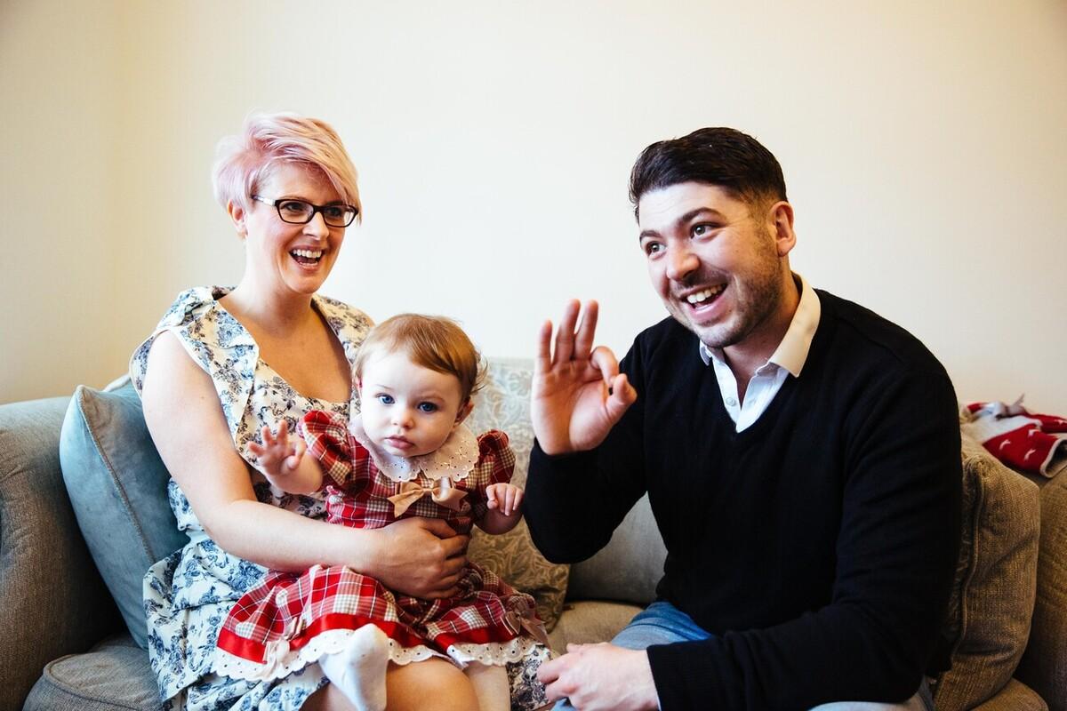 Primrose is reaching out while sitting on her mum's lap next to her dad signing "OK".