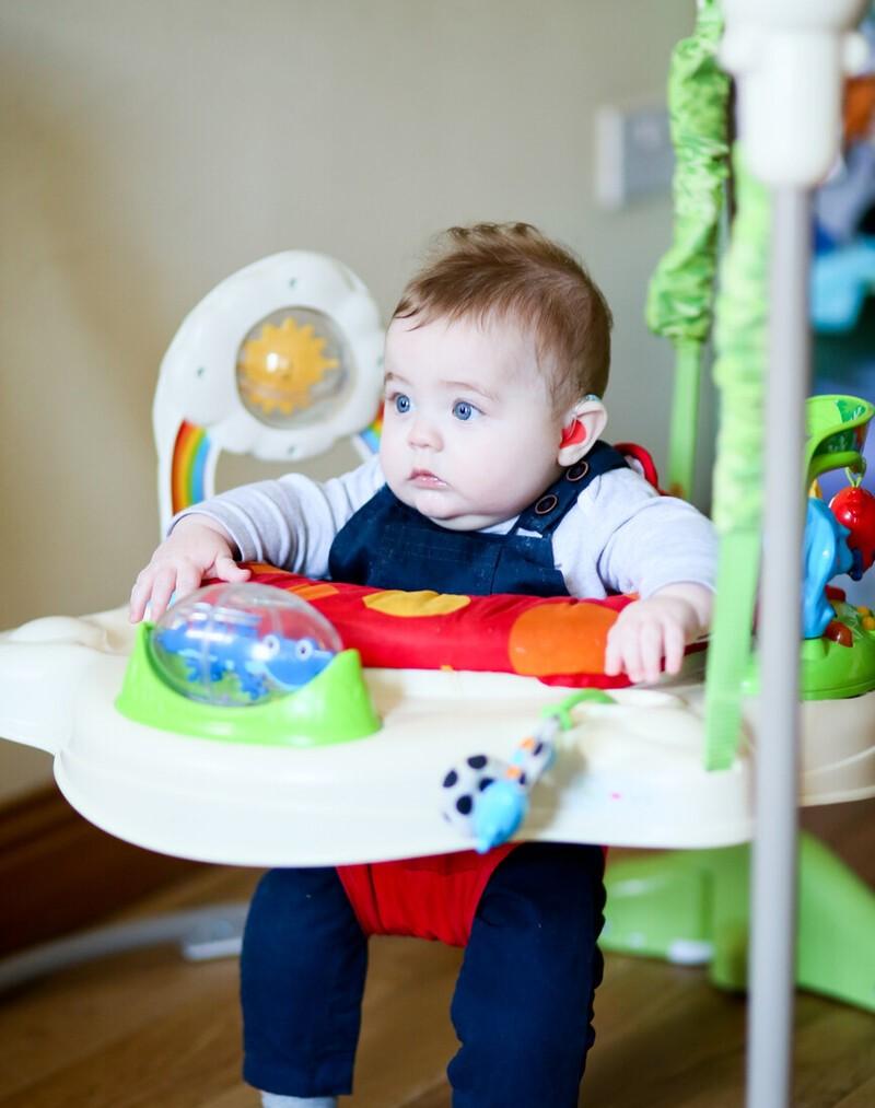 A baby is sitting in a bouncer. They are wearing a hearing aid.