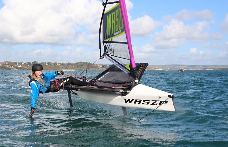 Girl in blue jacket leaning over and touching the water while in a Waszp sailing boat. 