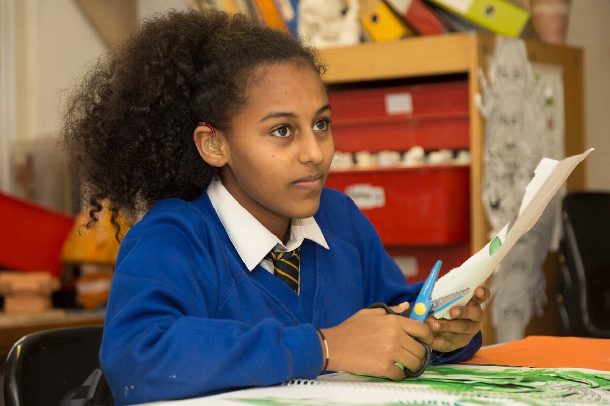A girl is sat in a classroom cutting some paper with scissors. She is wearing a hearing aid and has long dark curly hair in a ponytail. She is wearing a blue school uniform with a tie.
