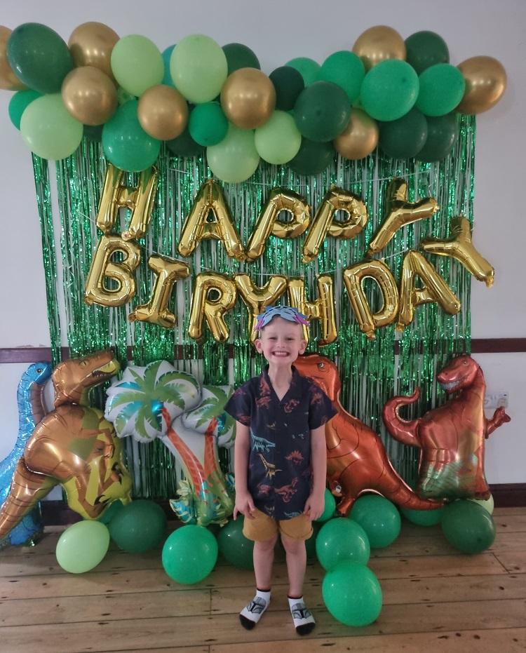 Kenzie smiling at the camera stood in front of a green and gold balloon and tinsel display, with blow up dinosaurs and 'Happy birthday' letter balloons.