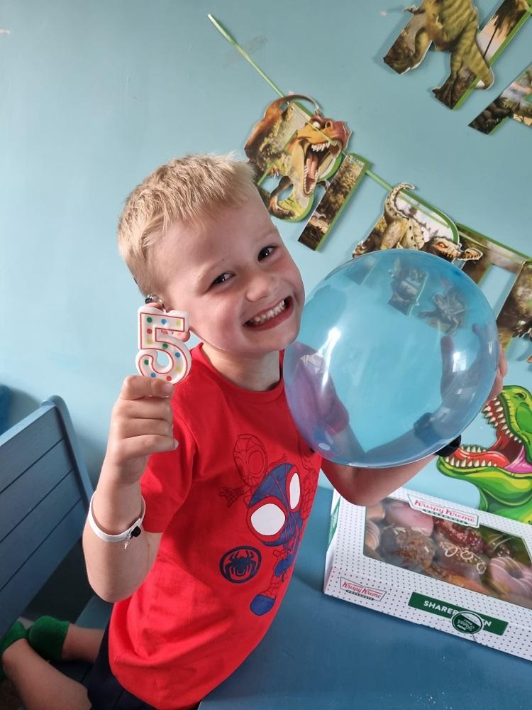 Kenzie smiling at the camera holding a 5 shaped birthday candle and a blue balloon.