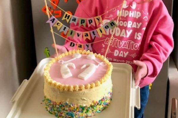 M wearing a pink jumper and holding her 'Happy Activation Day' cochlear implant cake.
