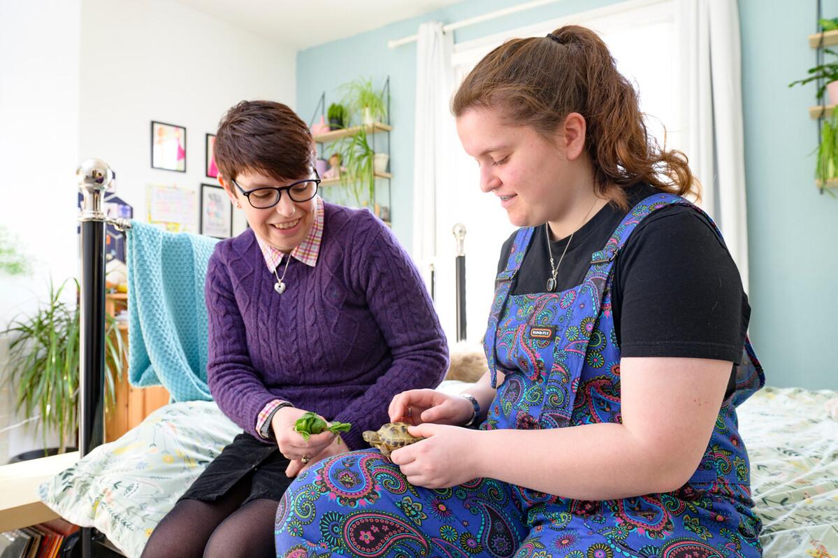 A teenage girl sits on her bed feeding a pet turtle with her mum beside her