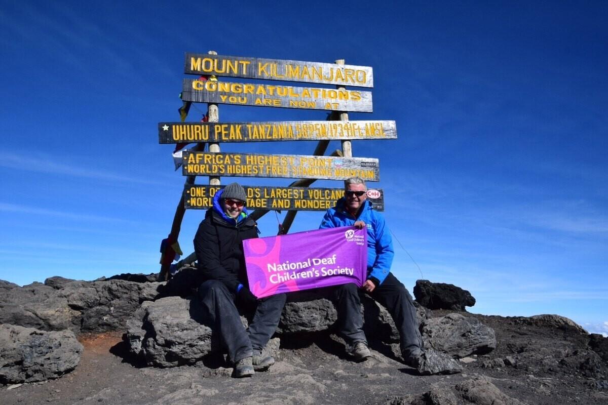 Two men sitting at Mount Kilimanjaro summit with a National Deaf Children's Society banner