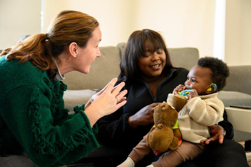 A woman looking at the young child who is sat on her lap while another woman is communicating with him