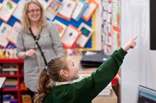 A girl with a hearing aid points at the whiteboard in front of a teacher.
