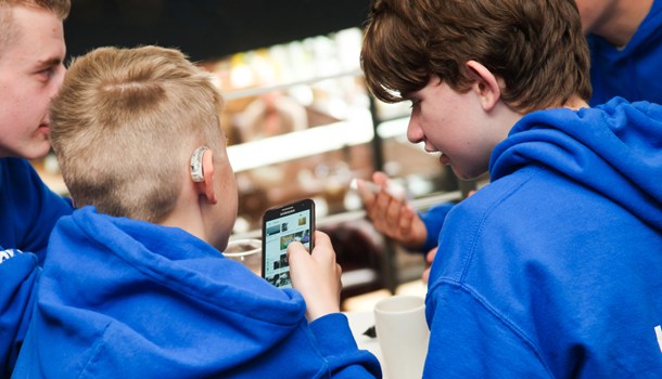 A boy with a hearing aid holds a phone while speaking to a group of friends.