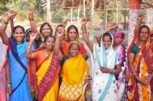 Group of women waving and smiling at the camera, they are wearing saris.
