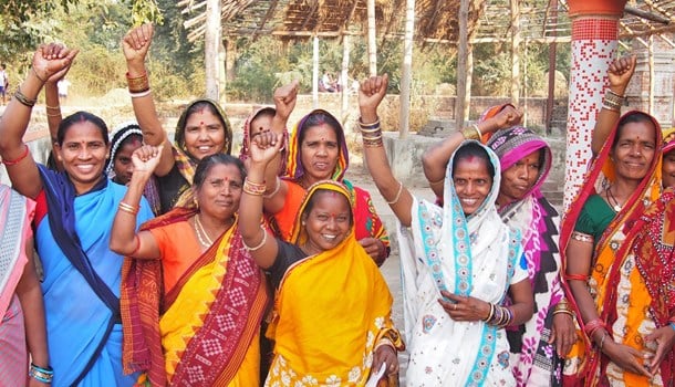Group of women waving and smiling at the camera, they are wearing saris.