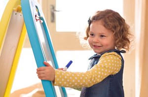 A smiling toddler draws on a whiteboard easel. 