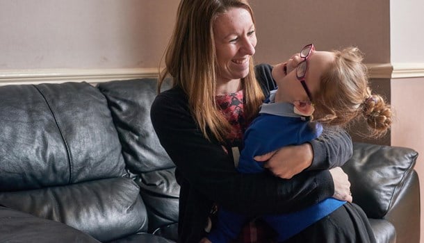 A smiling mum hugs her daughter who has cochlear implants and glasses on the sofa.