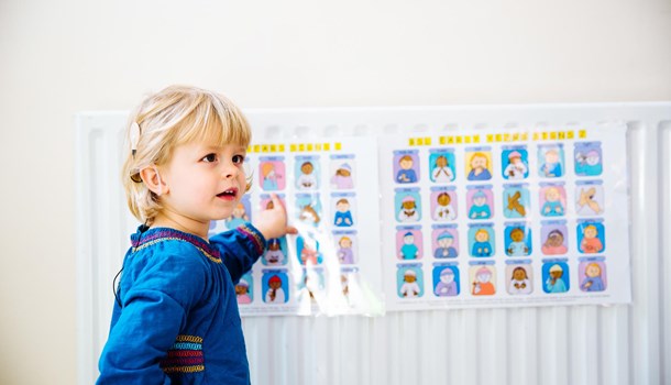 A young girl wearing a cochlear implant points to a poster with different BSL signs on it. 