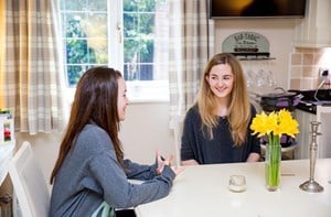 Two teenagers talk to each other at a kitchen table. 