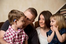 A mom and dad hold two young kids on their lap, the young boy is wearing hearing aids.