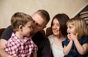A mom and dad hold two young children on their lap, the young boy is wearing hearing aids.