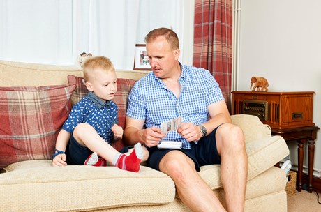 A dad shows his son, who has a bone anchored hearing aid (BAHA), learning cards while sitting on the sofa.