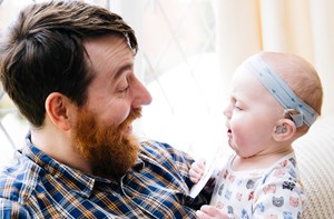 A dad smiles down at his baby who is wearing a hearing aid. 