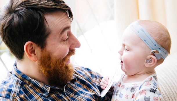 A dad smiles down at his baby who is wearing a hearing aid. 
