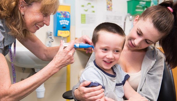 A smiling boy sits on his mum's lap while an audiologist examines his ear.