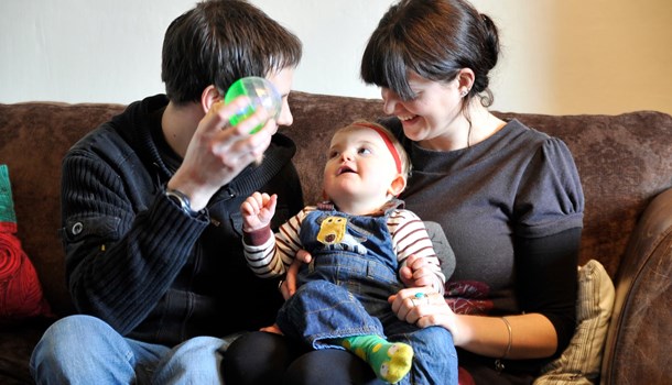 A mum and dad sit on the sofa holding a plastic toy ball up to their baby.