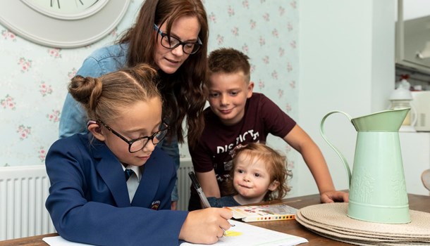 A girl wearing a hearing aids works on homework while her mum and two younger siblings look over her shoulder.