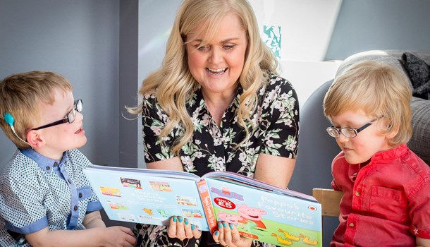 A mum reads a Peppa Pig picture book to her two young sons, one of whom is wearing a cochlear implant.