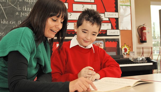 A boy with hearing aids sitting next to his teaching assistant looking at an exercise book.