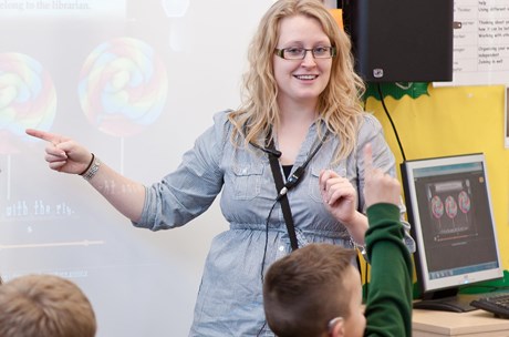 Young, smiling, female teacher wearing a radio aid in front of whiteboard in classroom and young child with hand up.